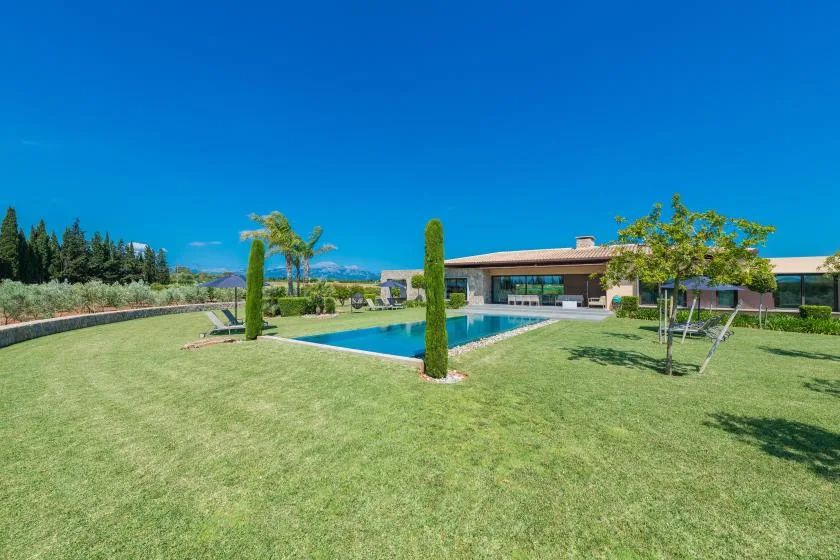 A modern house overlooks a rectangular swimming pool set in a broad lawn.