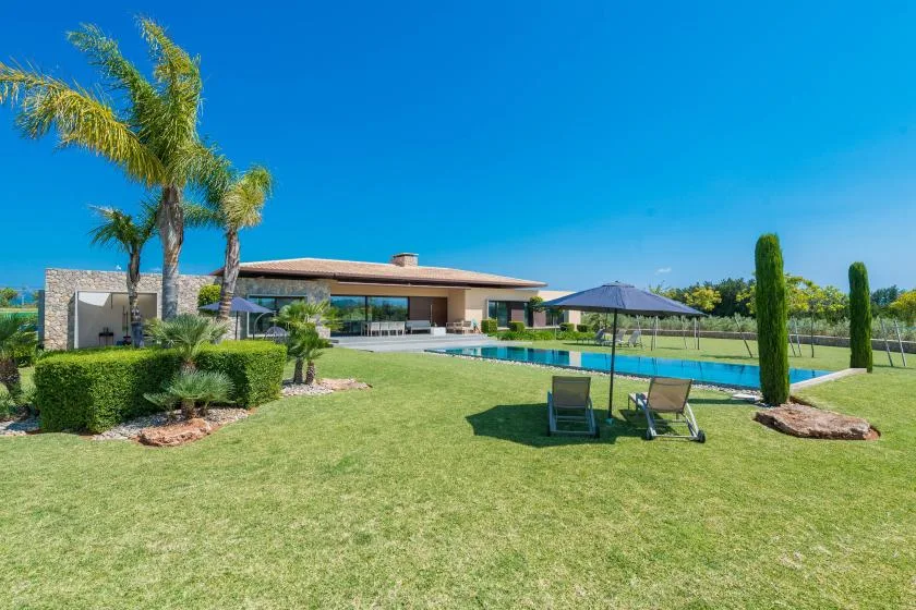 A modern house overlooks a lawn and swimming pool beneath a clear blue sky.