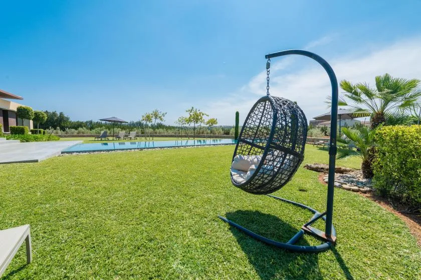 A grassy outdoor area features a hanging egg chair beside a swimming pool.