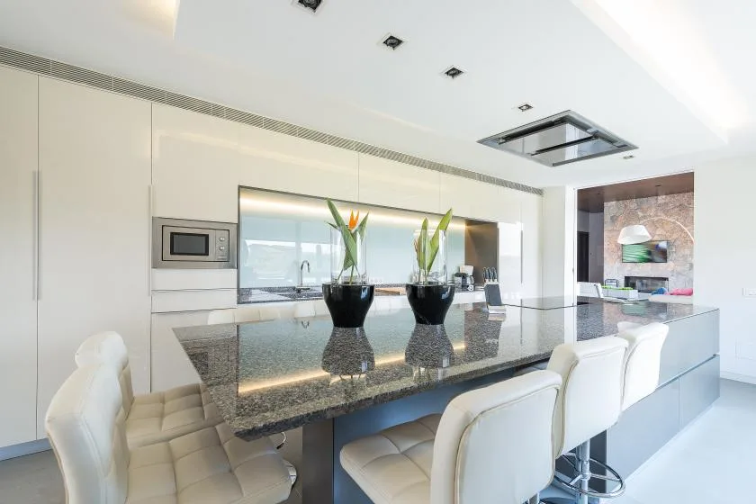 A modern kitchen with a dark island, white seating, and potted plants.