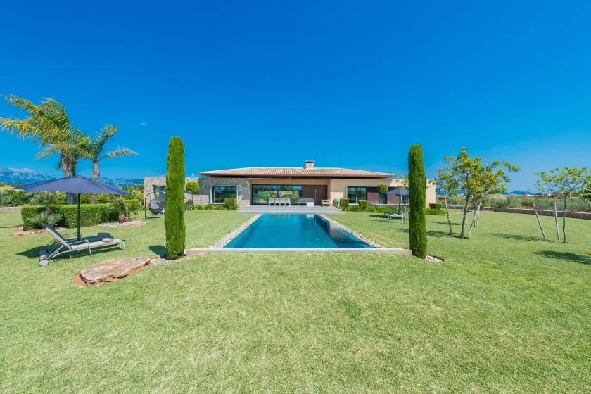 A modern house overlooks a rectangular swimming pool set in a broad green lawn.