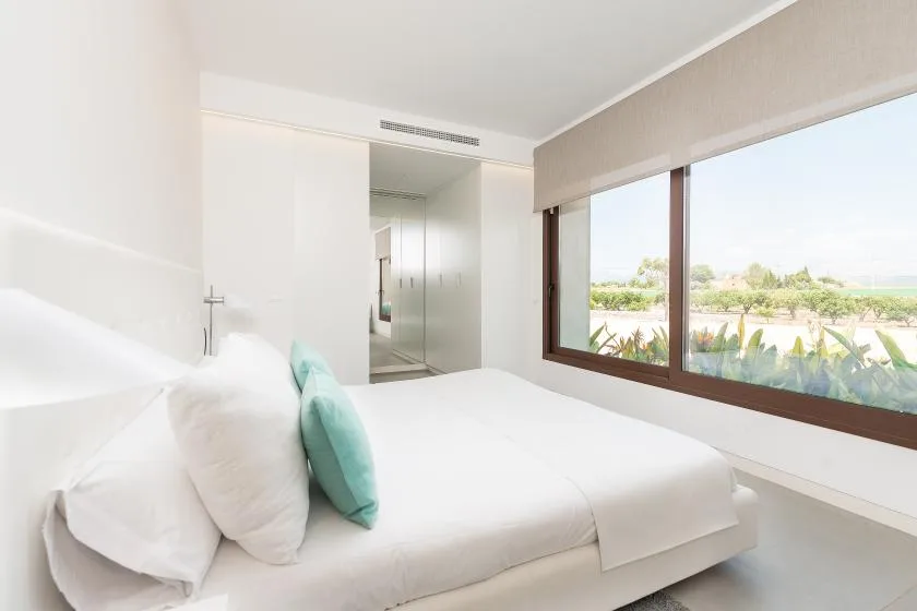 A bright bedroom with a white bed beside a large window.