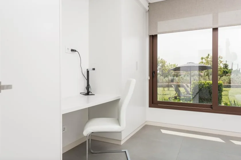 A bright workspace has a white desk and chair beside a large window overlooking greenery.