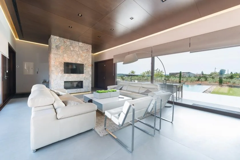 A modern living room with white seating, a stone fireplace, and large windows overlooking a pool.