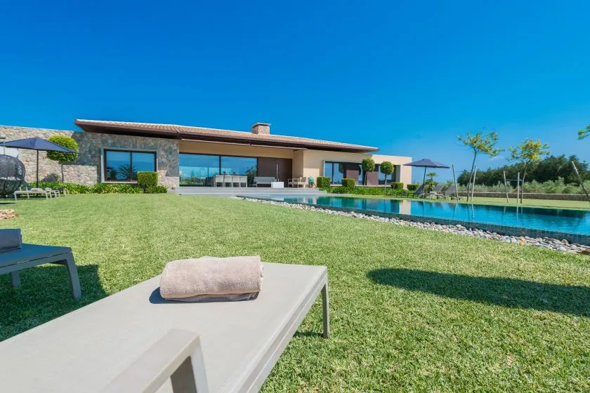 A modern house overlooks a swimming pool and lawn beneath a clear blue sky.