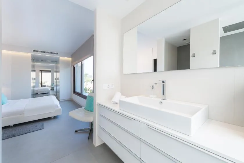 A bright bedroom with a bed beside a long white vanity and mirror.