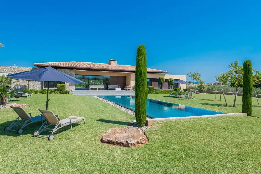A modern house overlooks a swimming pool surrounded by a lawn.