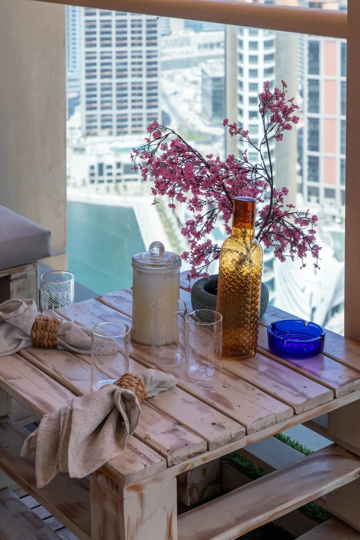 Una mesa junto a una ventana tiene cristalería, una vela y ramas en flor.