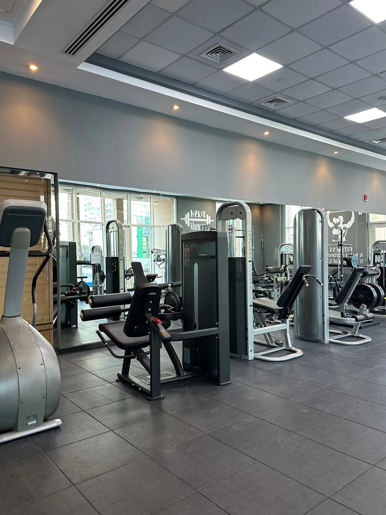 Un gimnasio con máquinas de ejercicio y grandes ventanas a lo largo de una pared.