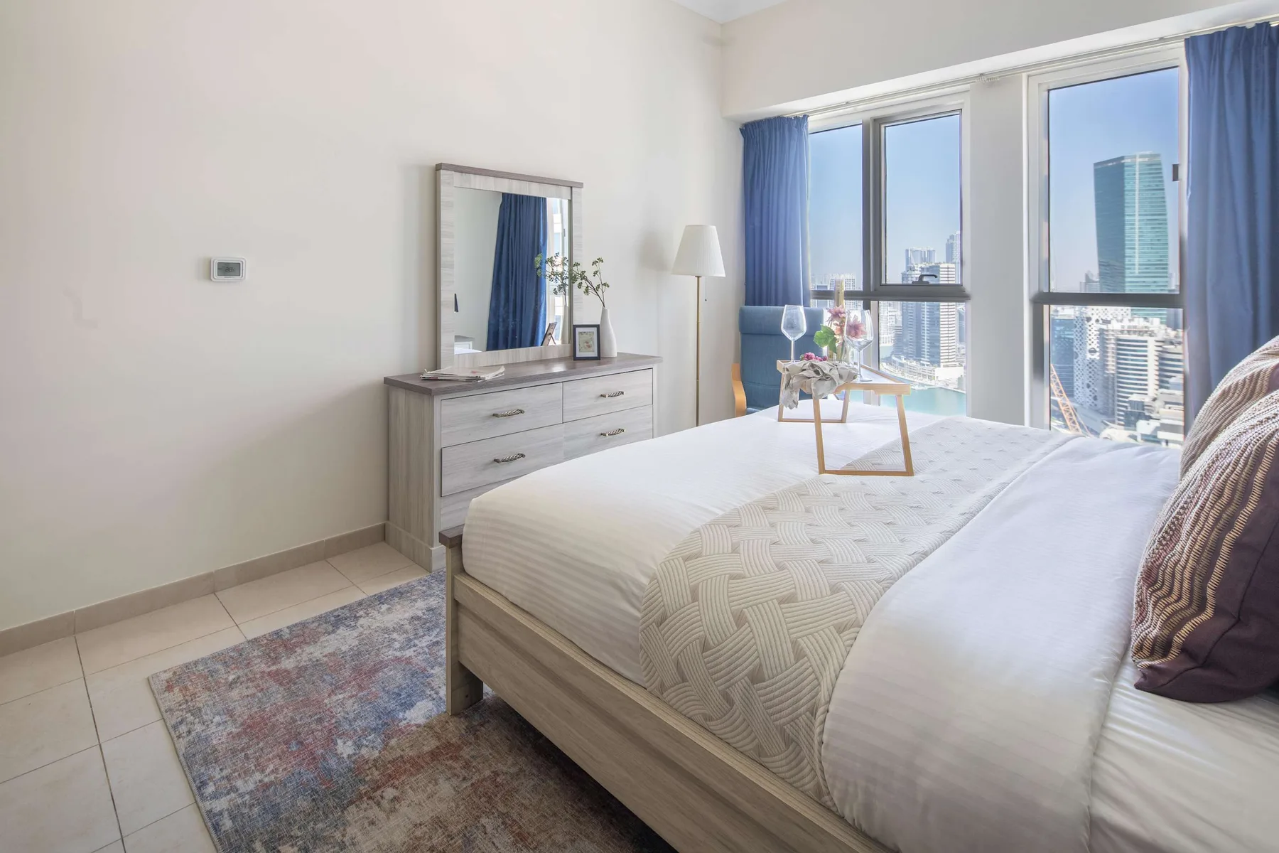 Un dormitorio luminoso con una cama tendida junto a ventanas altas con vistas a la ciudad.
