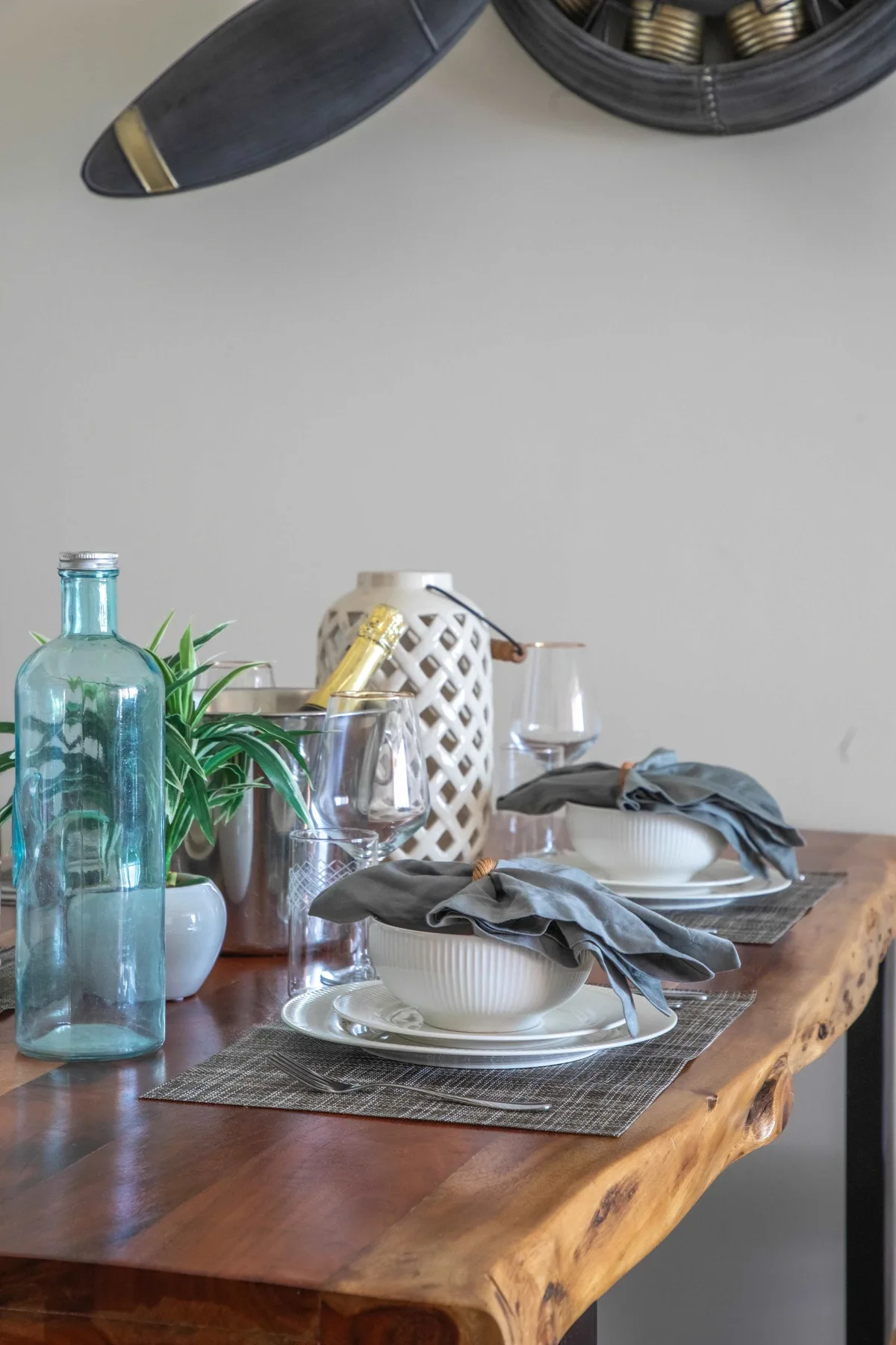 Una mesa de comedor está puesta con platos, cuencos, cristalería y un jarrón con plantas.