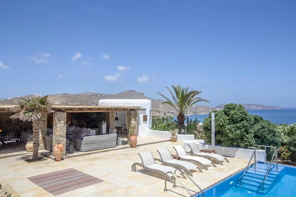 A sunny pool terrace with lounge seating, palm trees, and a view of the sea.