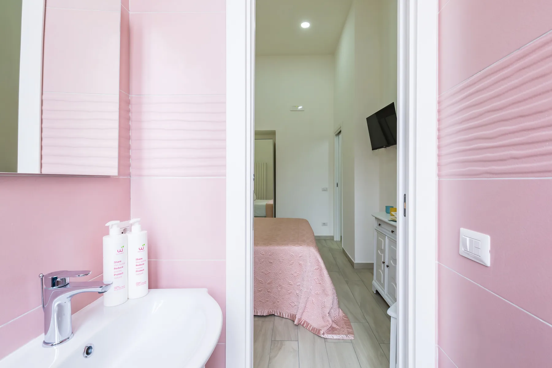 A pink-tiled bathroom opens onto a bedroom with a bed and wall-mounted television.
