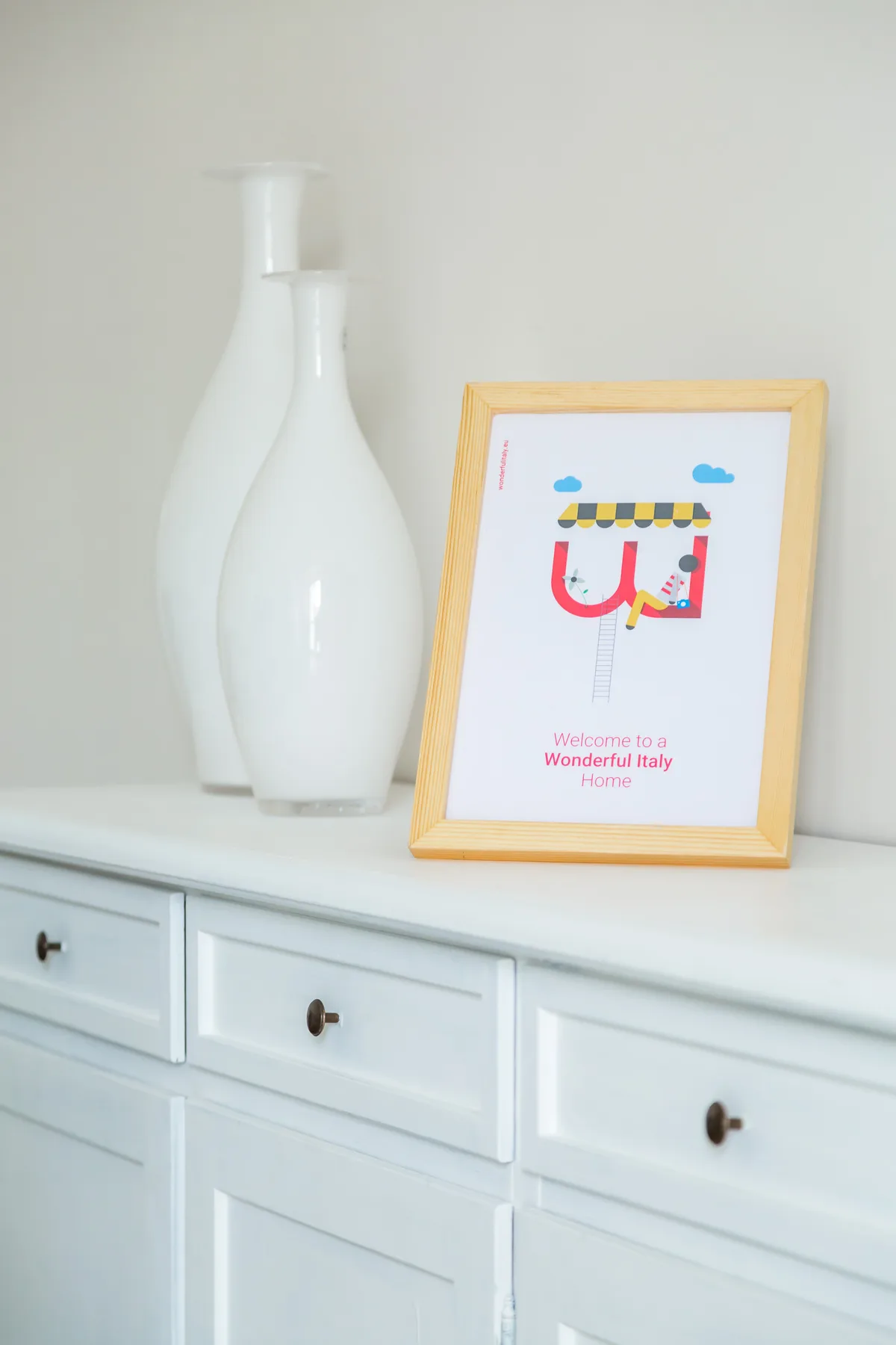 A white dresser displays two white vases and a framed colorful illustration.