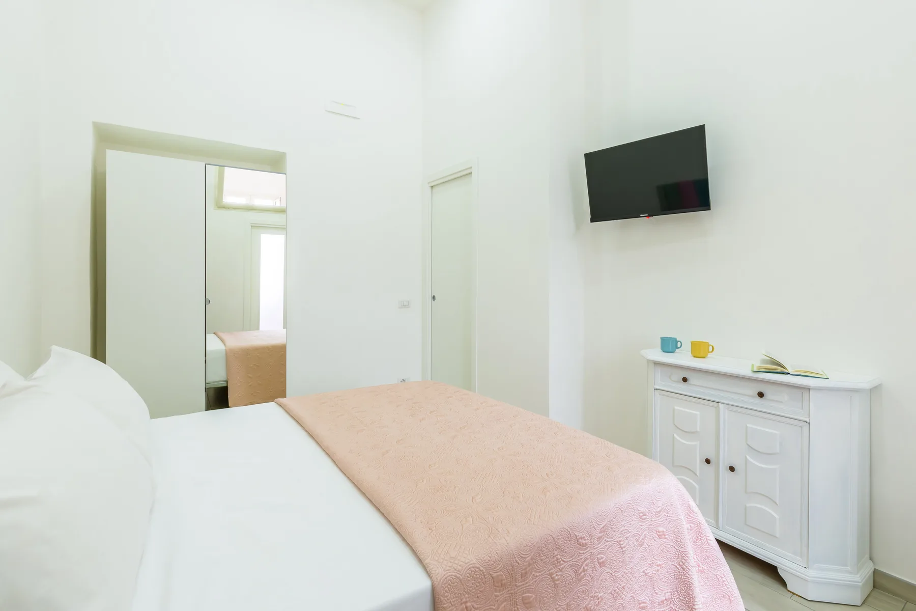 A bright bedroom with a bed, wall-mounted television, and white cabinet.