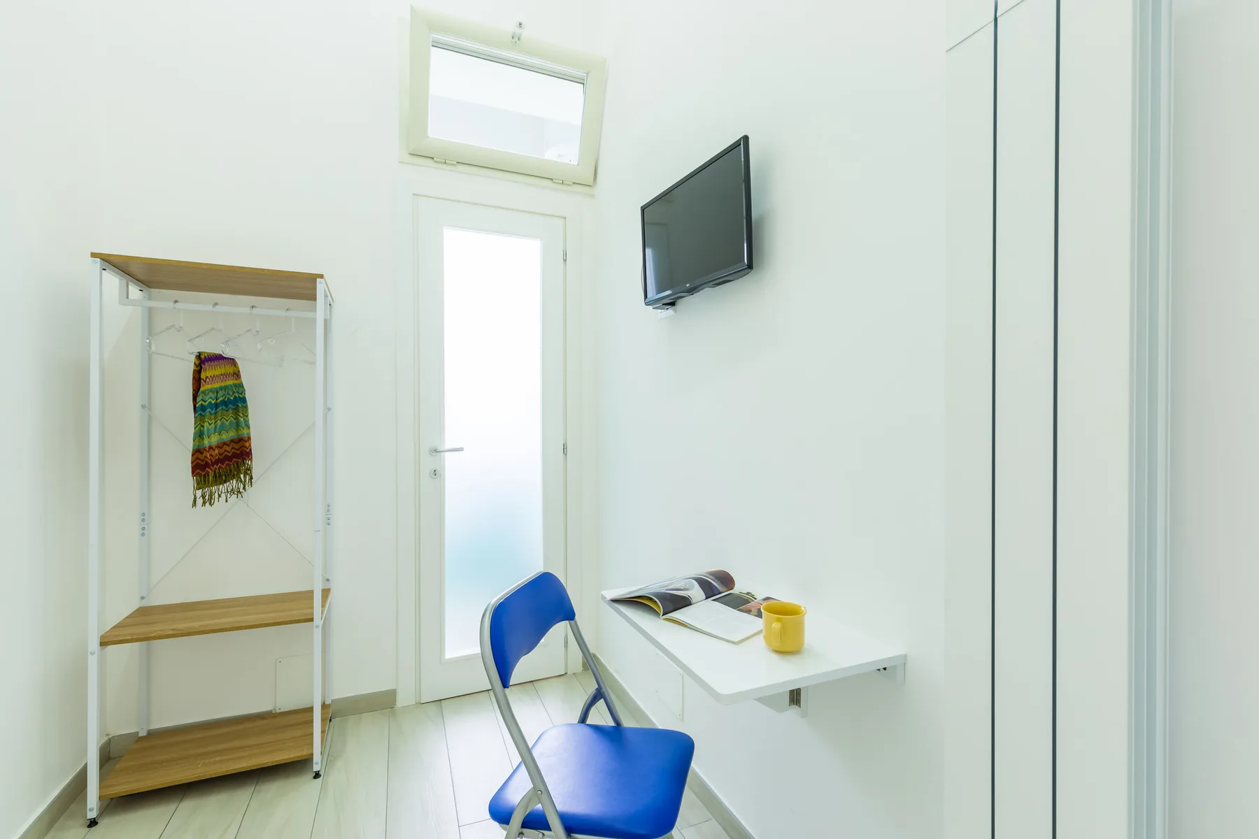 A bright room has a blue chair beside a small desk and wall-mounted television.