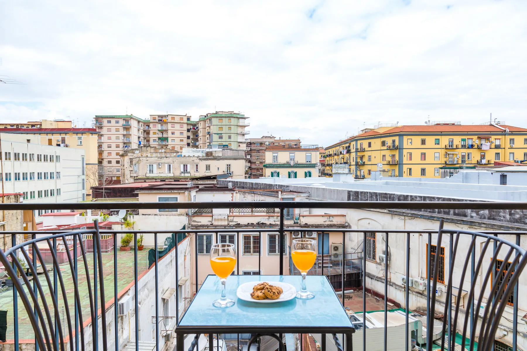 A balcony dining area overlooks a city neighborhood, with drinks and food on the table.