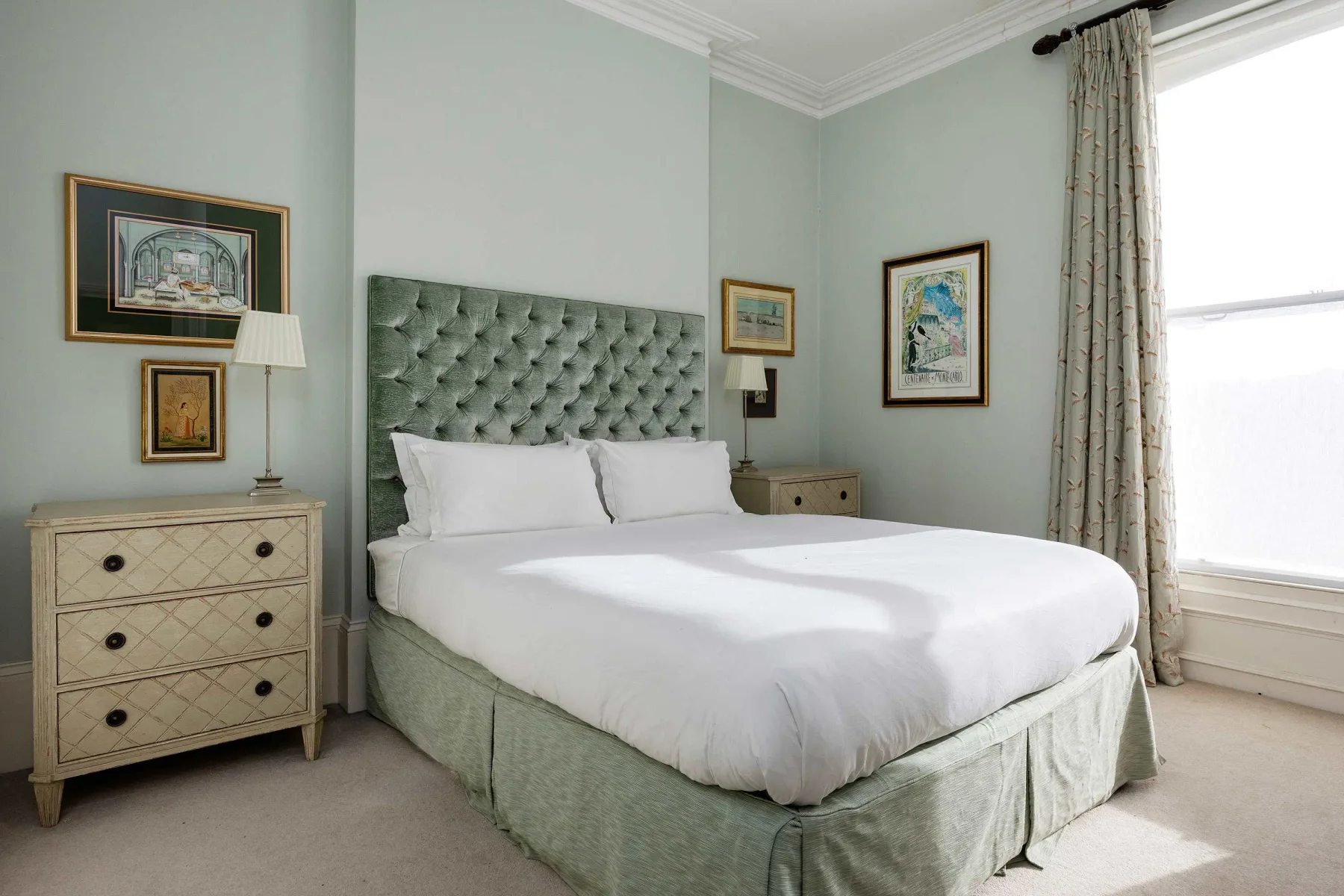 A bedroom with a large bed, pale green upholstered headboard, and a bright window.