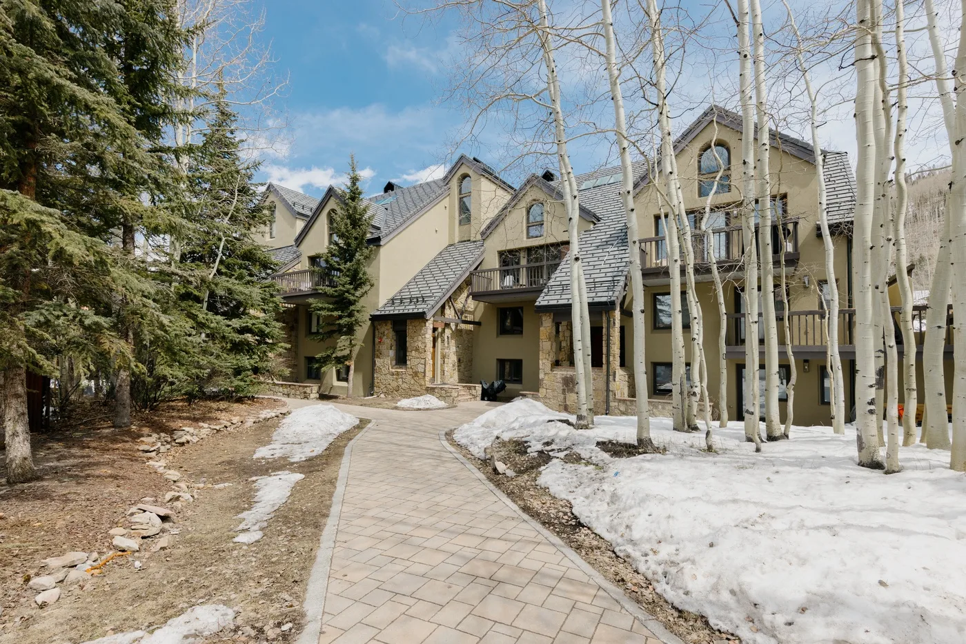 A paved walkway leads toward a large lodge-style building among trees and patches of snow.