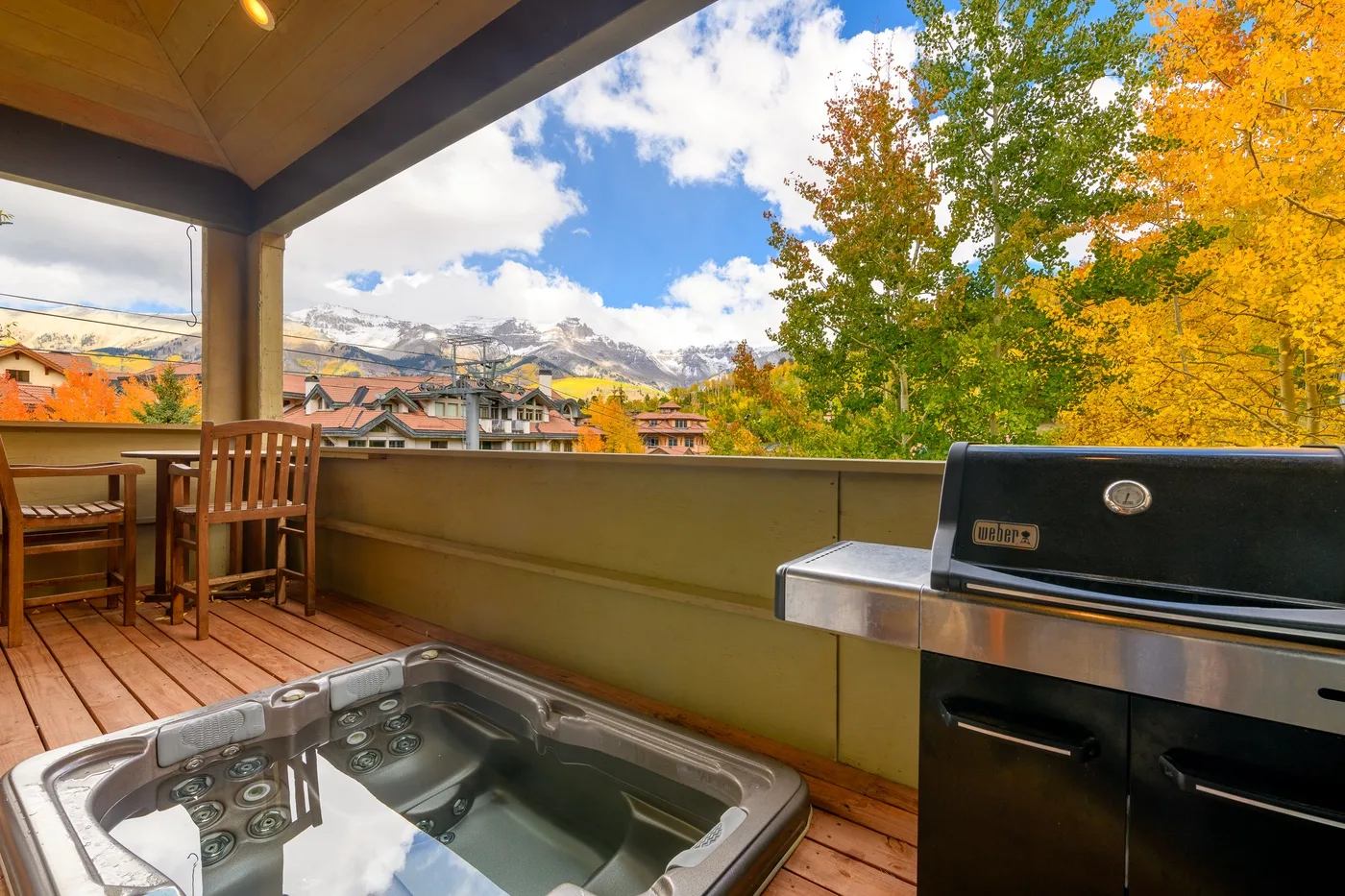 A covered deck with a hot tub and grill overlooks autumn trees and distant mountains.