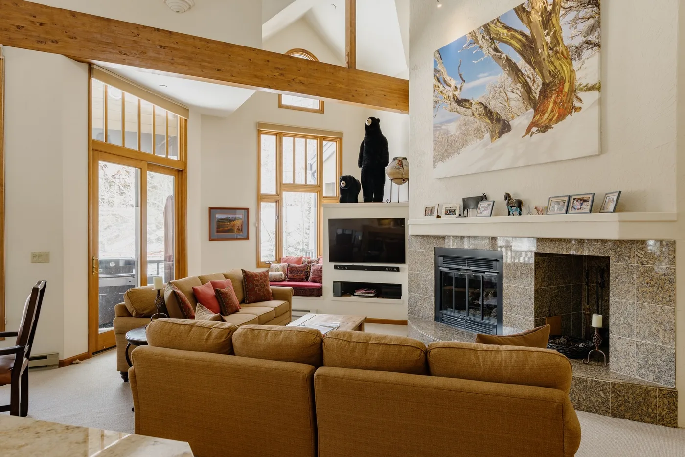 A bright living room with tan seating, tall windows, and a stone fireplace.
