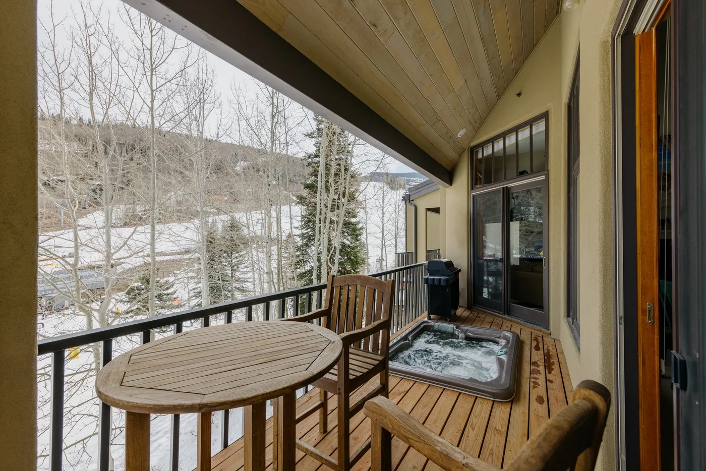 A covered wooden deck has seating and a hot tub beside large windows.