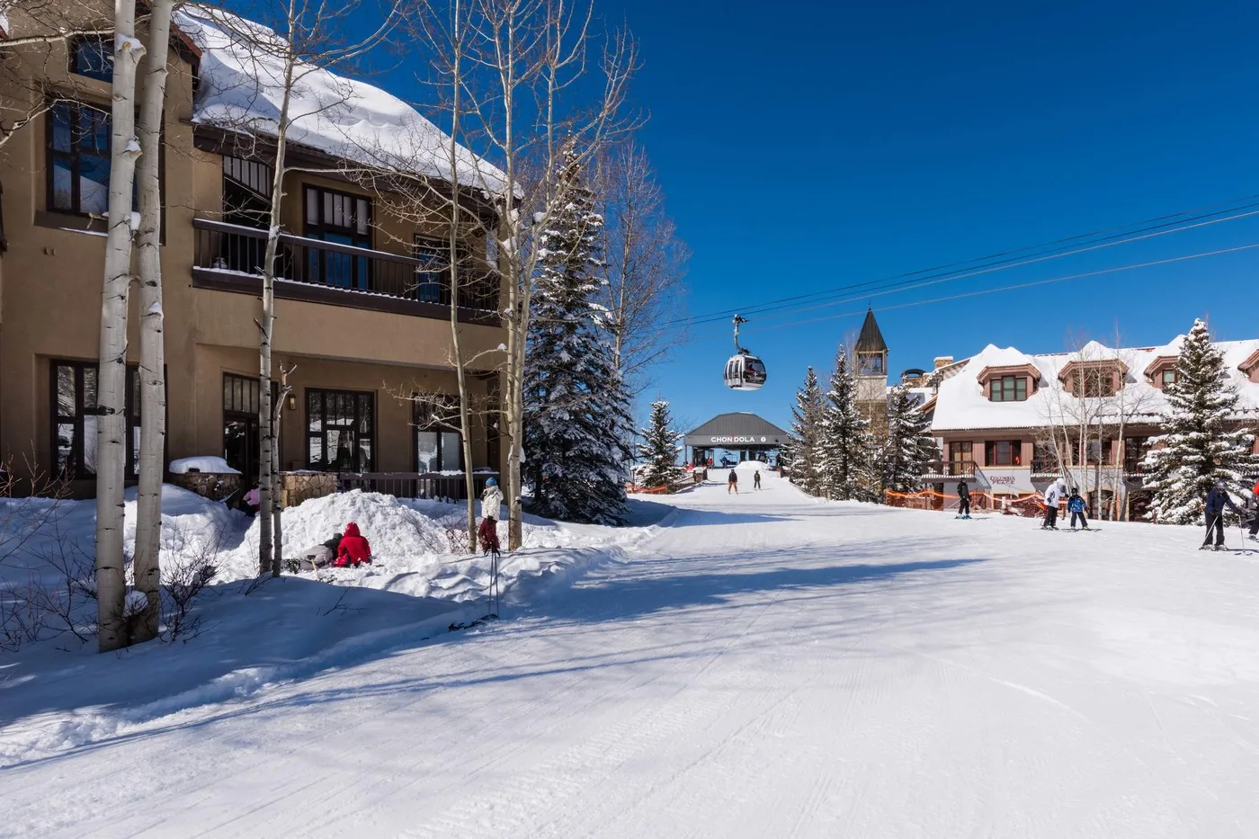 Snow-covered resort buildings and evergreen trees border a ski slope beneath a clear blue sky.