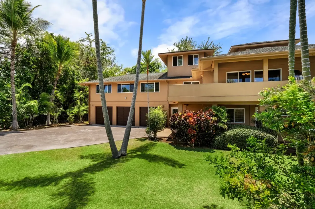 Una casa grande de color beige se alza detrás de un césped y vegetación tropical.