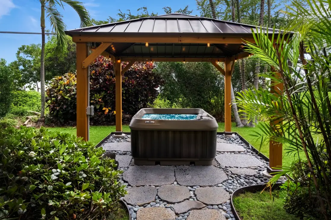 Una bañera de hidromasaje al aire libre está bajo una pérgola de madera, rodeada de vegetación tropical.
