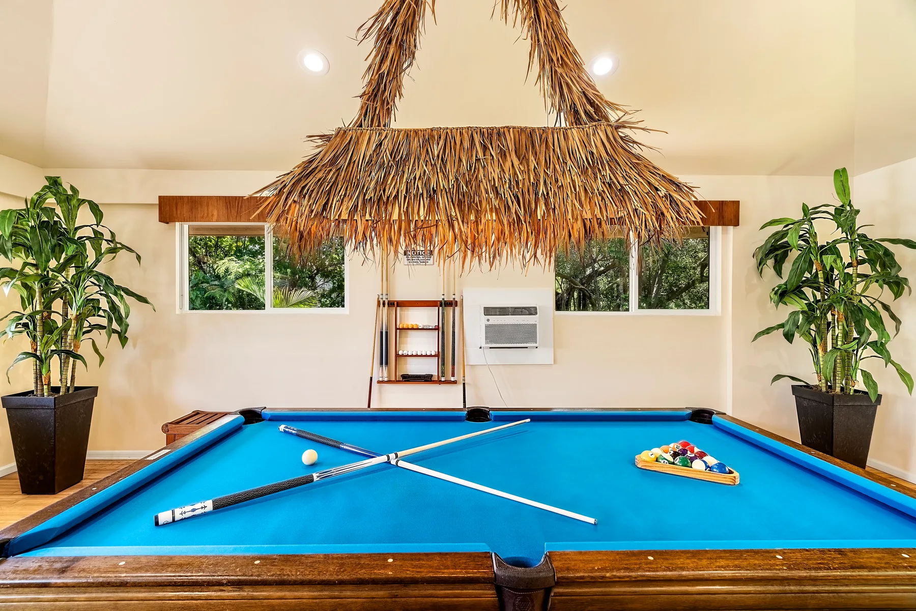 Una mesa de billar está bajo una gran lámpara colgante de paja, con plantas junto a las ventanas.