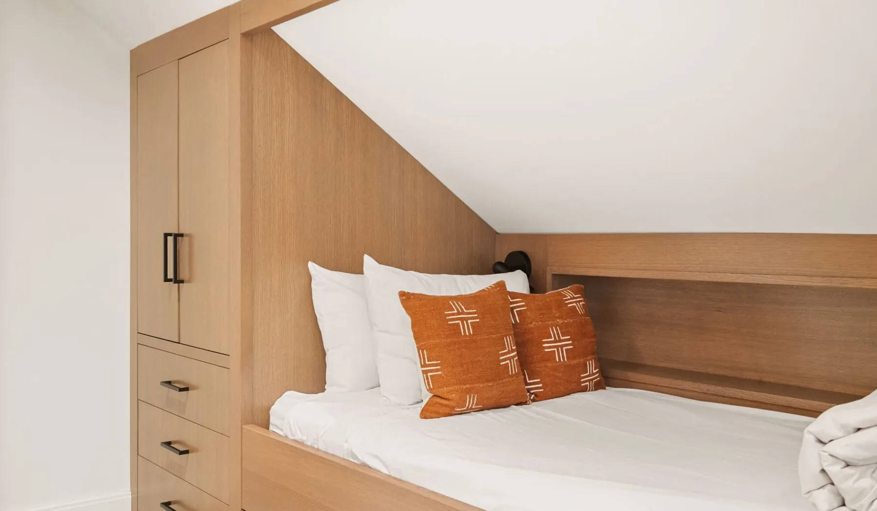 Dormitorio con una cama blanca y cojines decorativos color óxido bajo un techo inclinado.