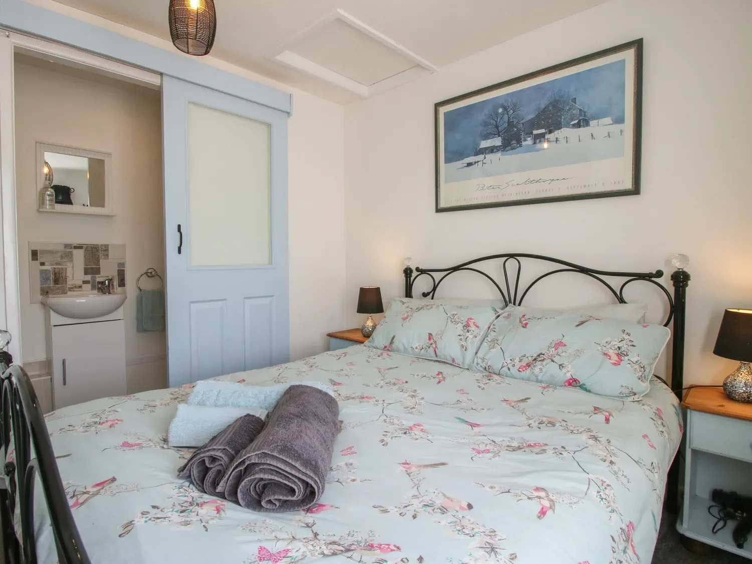 Un dormitorio con una cama con ropa de cama de flores y toallas encima, una mesita de noche con una lámpara y una puerta corredera que da a un baño pequeño en The Old Beams, en Pont y Forwyn, Llansantffraidd-ym-Mechain, cerca de Welshpool.