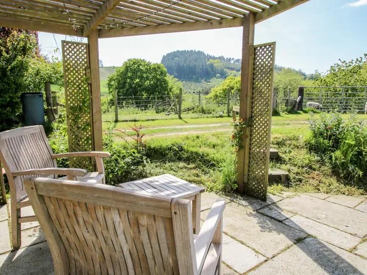 Un patio cubierto con asientos de madera da a un jardín verde y colinas lejanas.