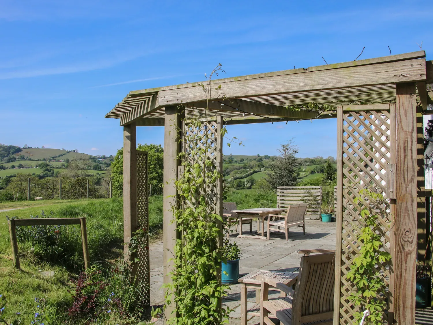 Una pérgola de madera con plantas trepadoras cubre asientos al aire libre en un patio pavimentado con vistas a colinas verdes.