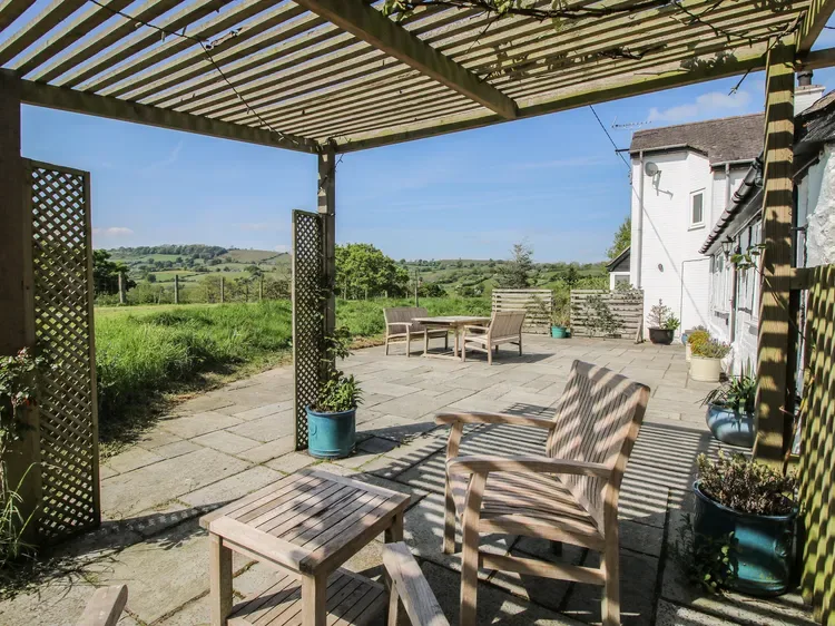 Un patio pavimentado bajo una pérgola tiene asientos al aire libre con vistas a campos verdes.