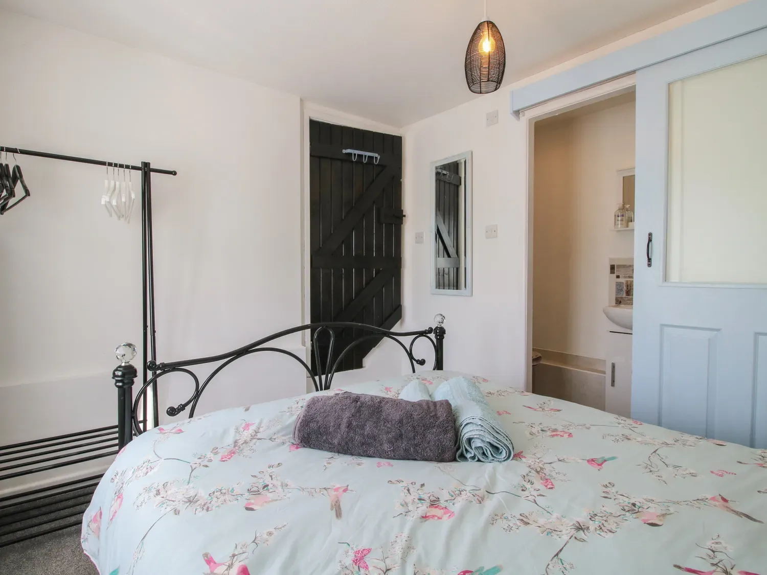 Un dormitorio con una colcha de flores, una estructura de cama de metal negro, toallas sobre la cama, un perchero negro, una puerta de madera negra, un espejo en la pared y una puerta corredera que da a un baño en The Old Beams, en Pont y Forwyn, Llansantffraidd-ym-Mechain, cerca de Welshpool.