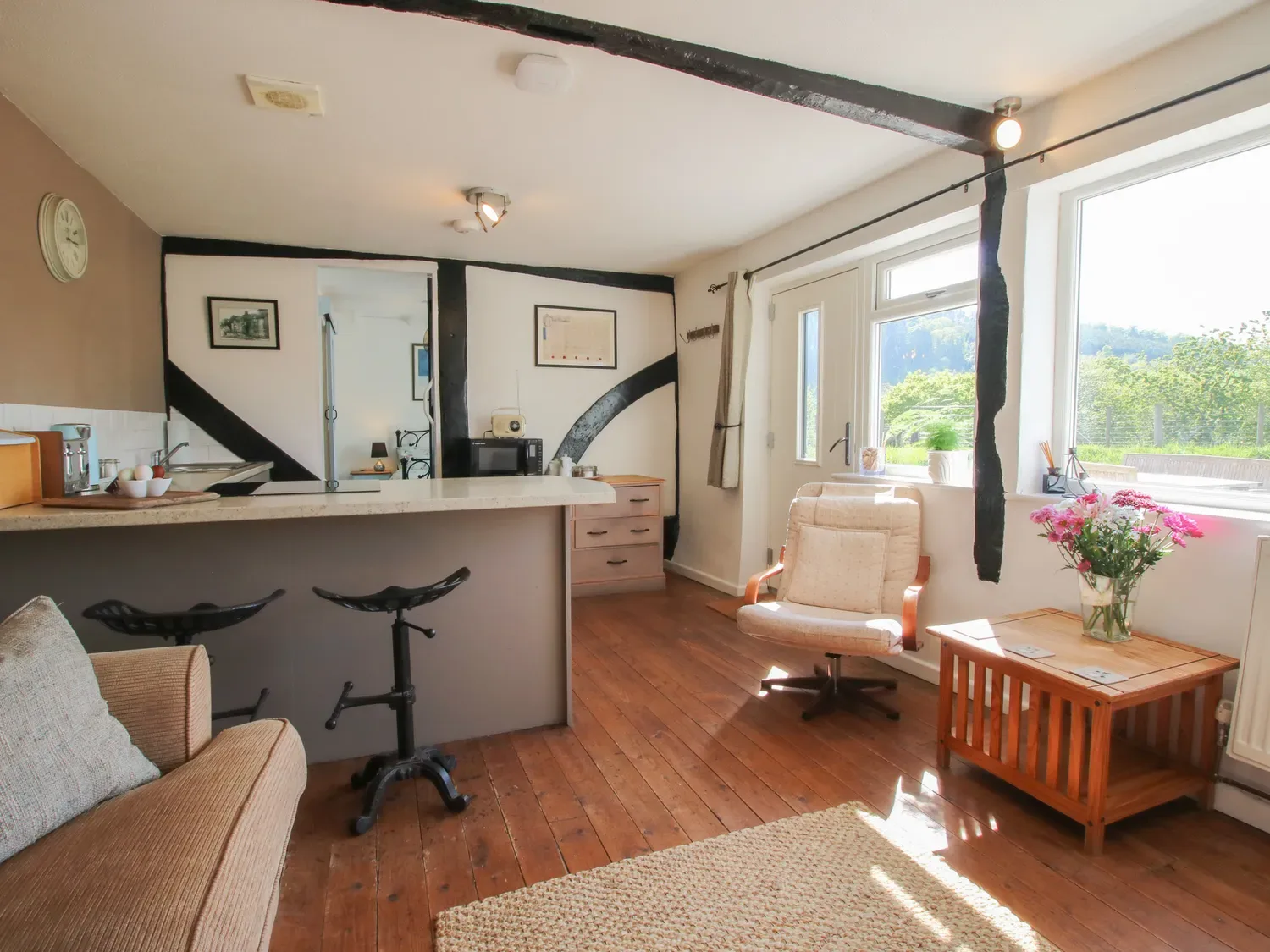 Una habitación con taburetes junto a una encimera, un sillón, una mesa auxiliar de madera con flores y ventanas grandes en The Old Beams, en Pont y Forwyn, Llansantffraidd-ym-Mechain, cerca de Welshpool.