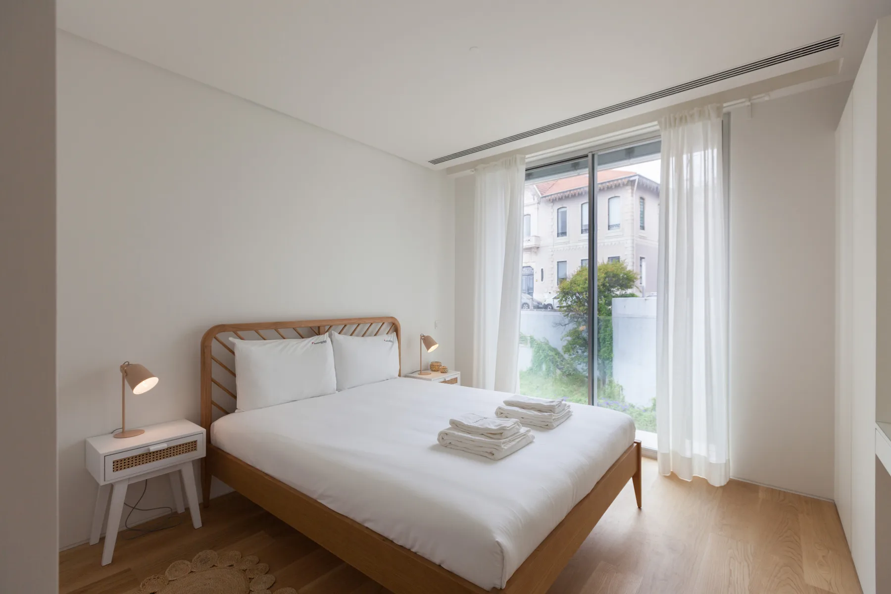 A bright bedroom with a neatly made bed beside a large curtained window.