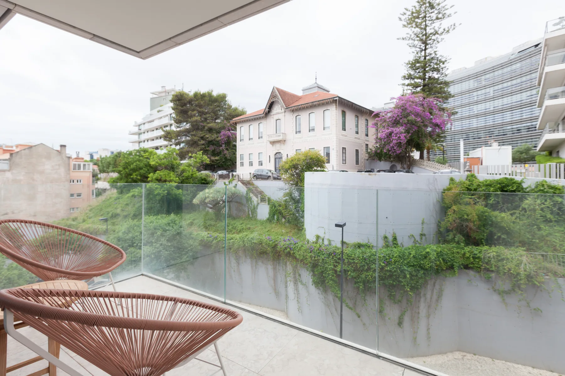 A balcony seating area overlooks a garden and nearby buildings.