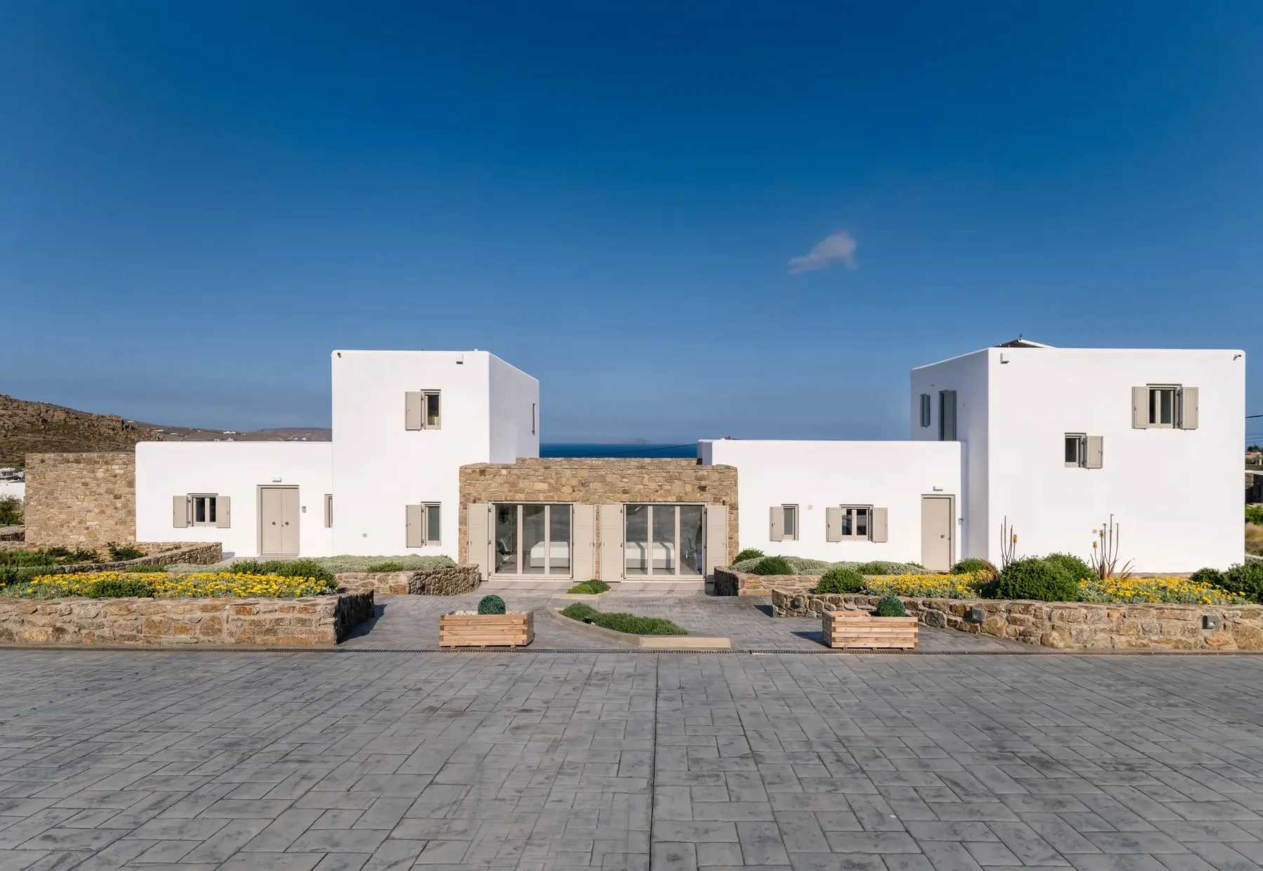 A modern white villa stands behind paved courtyard space and landscaped planters.