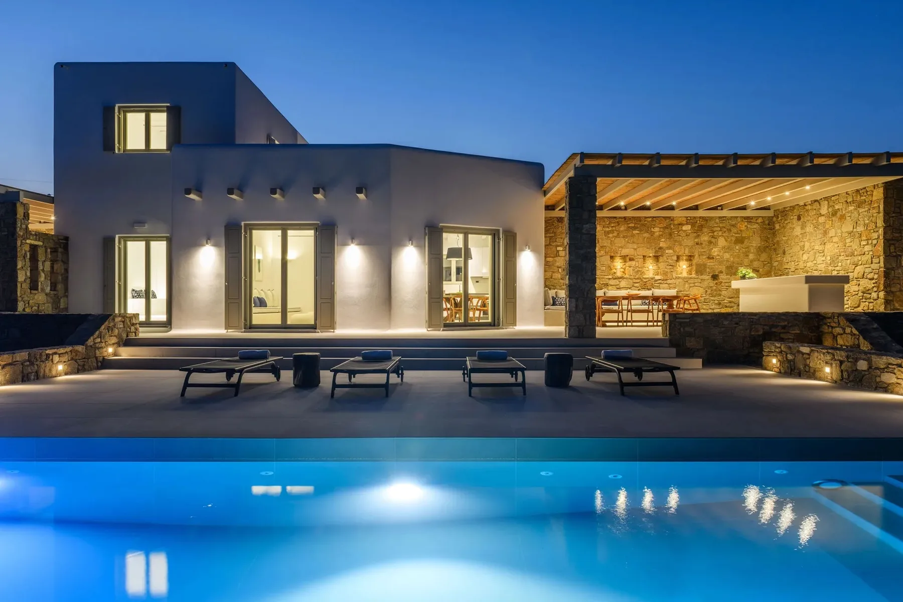 A modern villa exterior opens onto a lit swimming pool and covered outdoor seating area.