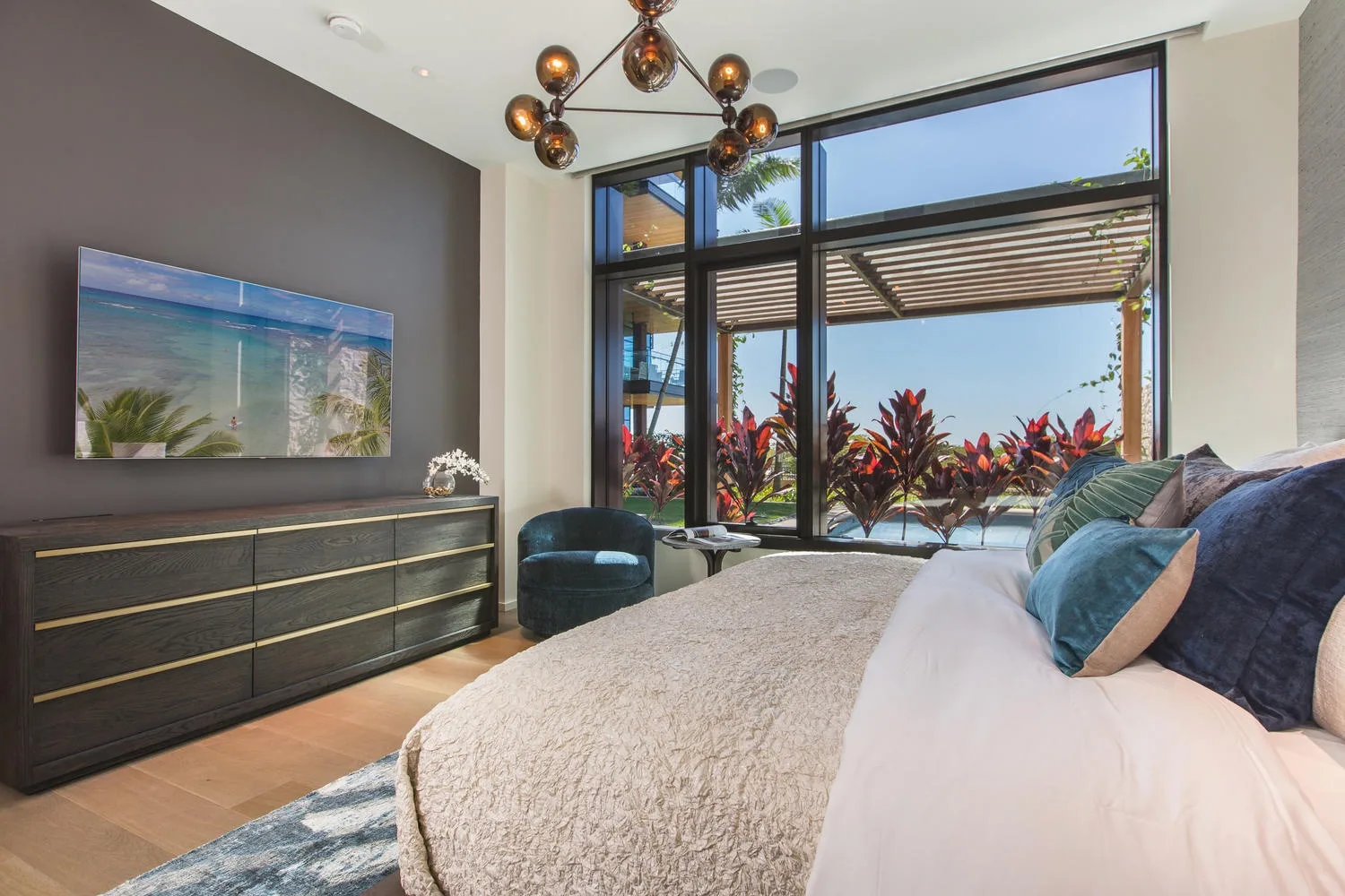 A bedroom with a large bed beside broad windows overlooking leafy plants.