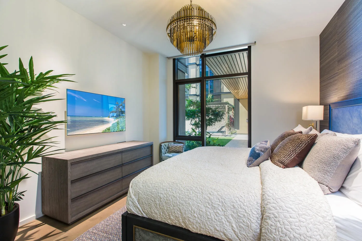 A bright bedroom with a bed, large window, and leafy plant.