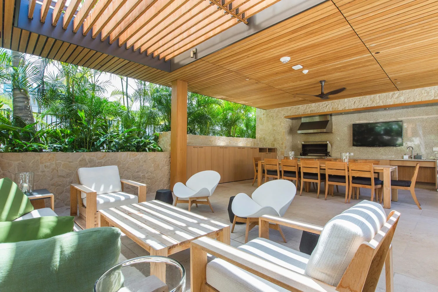 A covered patio lounge and dining area with seating, a long table, and greenery.
