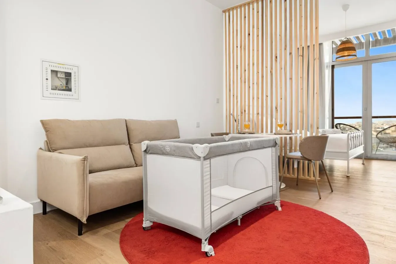 A bright living area with a beige sofa and a white playpen on a red rug.