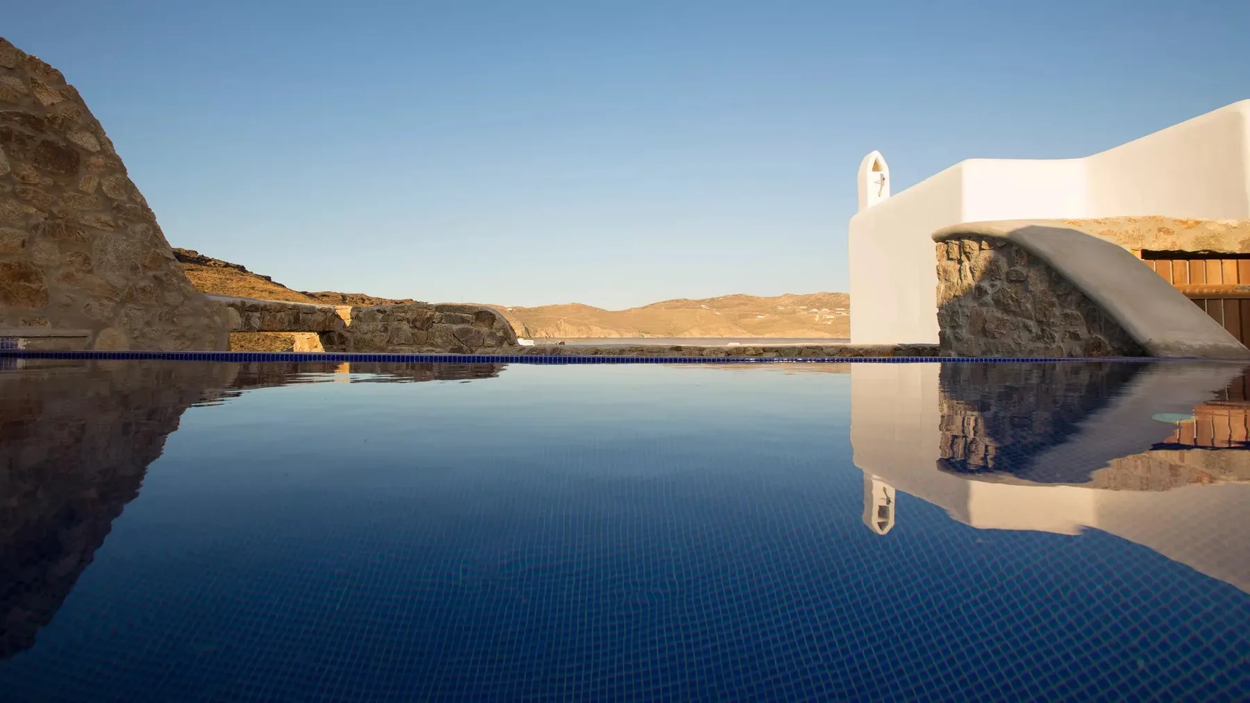A swimming pool reflects a white building and rocky hills.