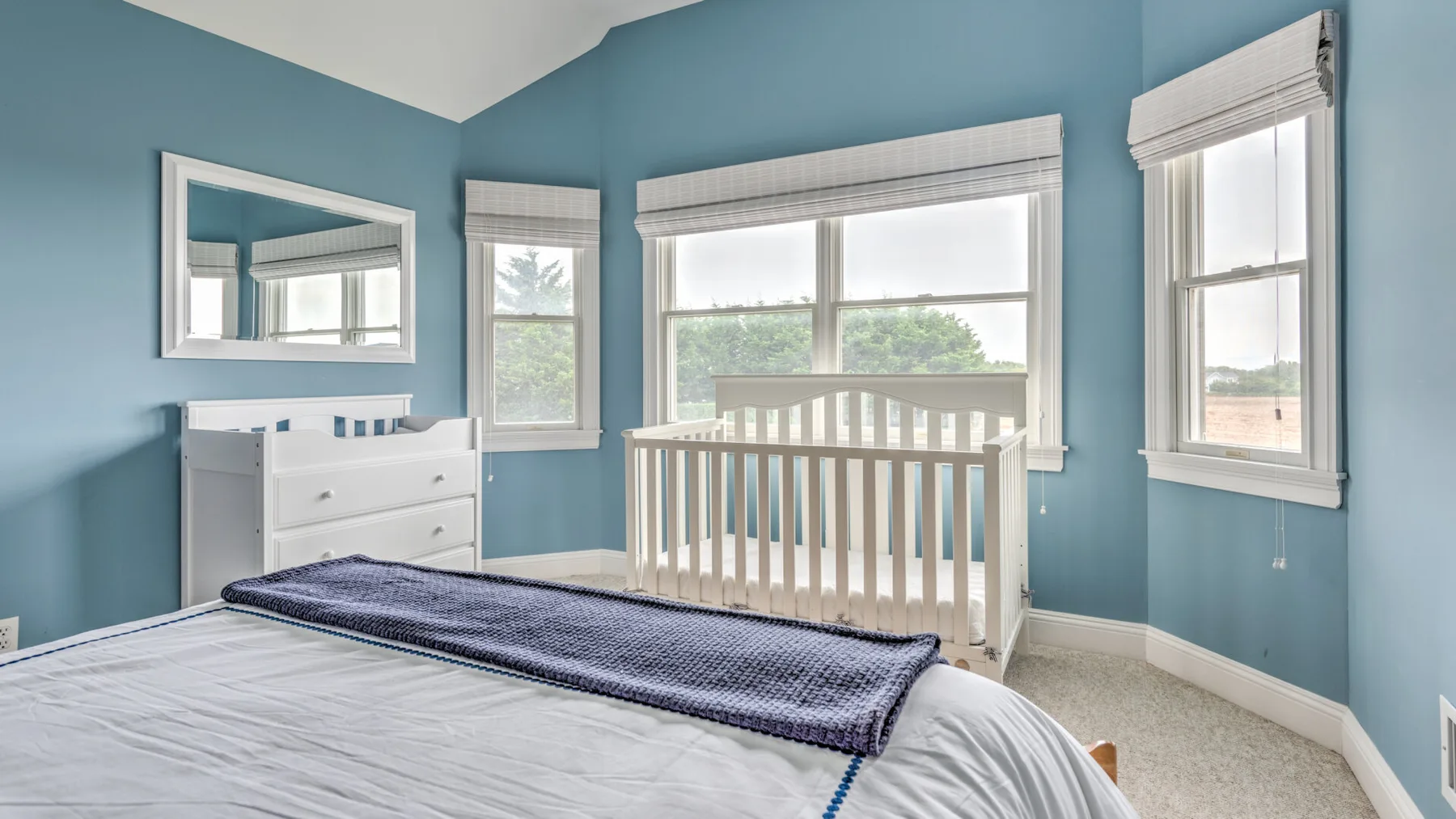Un dormitorio con una cuna blanca, una cómoda y ventanas grandes.