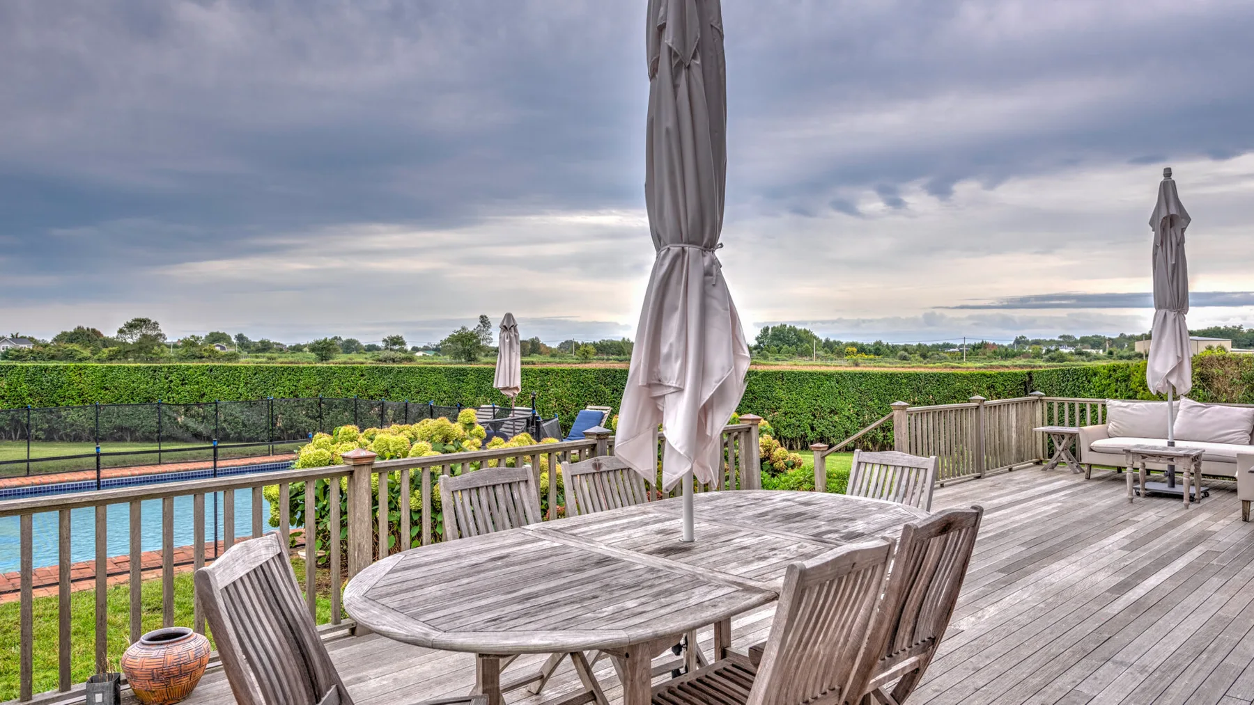 Una terraza exterior de madera tiene una mesa de comedor y asientos junto a una piscina y campos verdes.