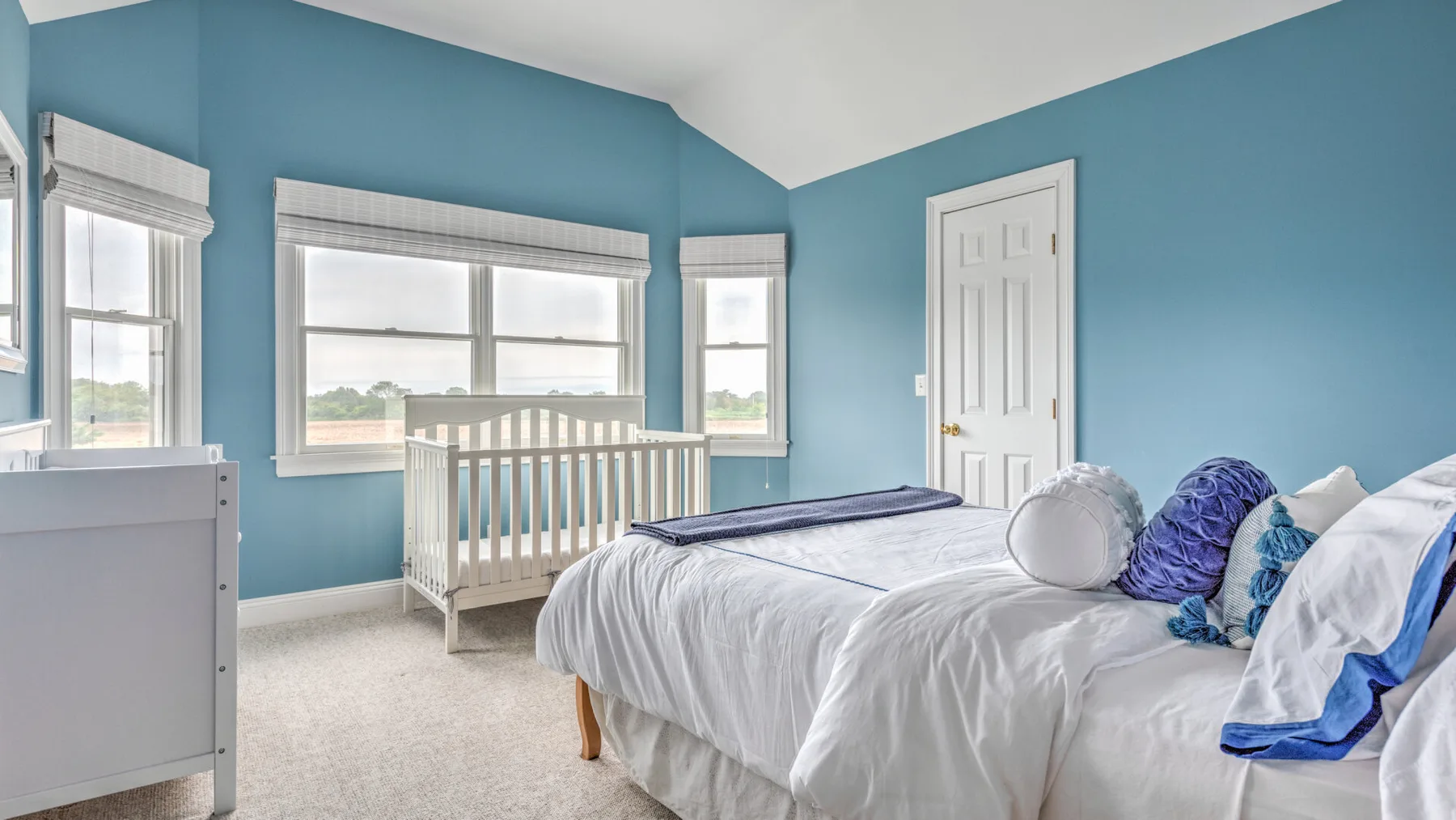 Un dormitorio luminoso con una cama, una cuna y ventanas con vistas a un paisaje verde.