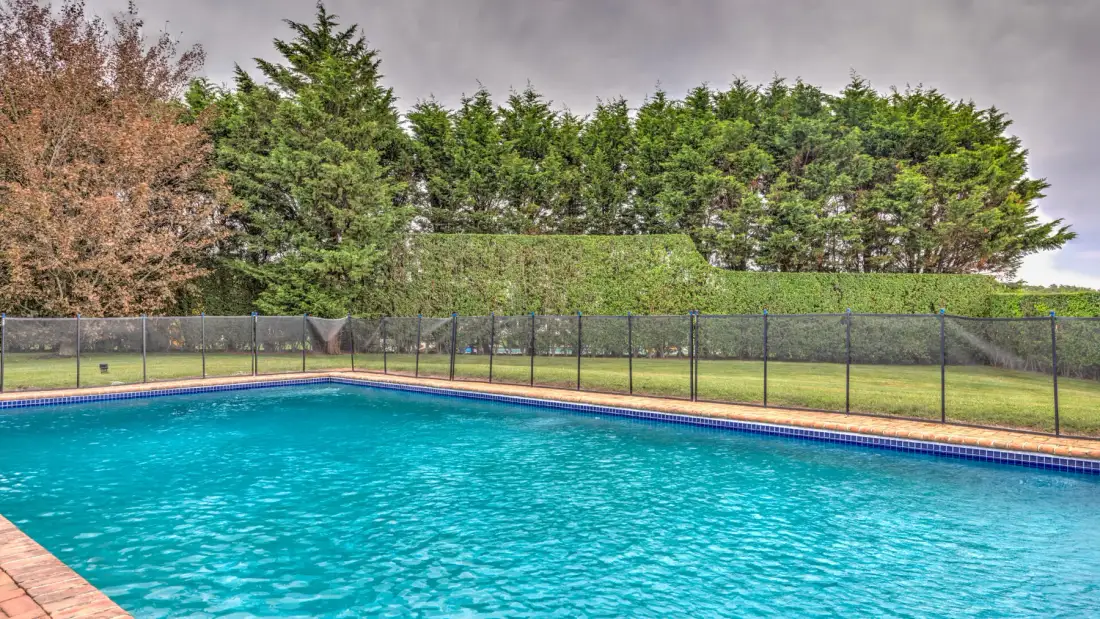 An outdoor swimming pool sits beside a fenced lawn and tall trees.